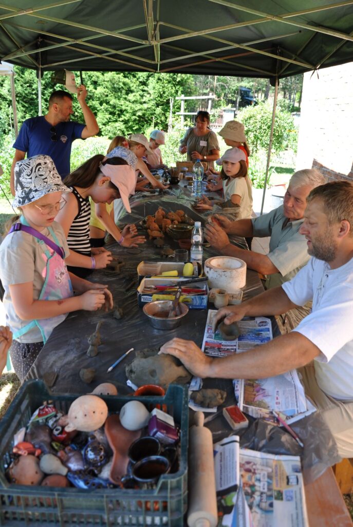 Grupa ludzi siedzi wokół stołu, dorośli i dzieci, wykonują różne przedmioty z gliny.
