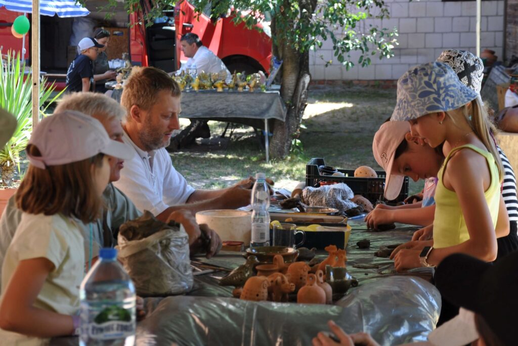 Grupa ludzi, dorośli i dzieci siedzą przy stole wykonując ręcznie przedmioty z gliny.