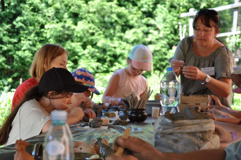 Grupa ludzi, dwie kobiety i troje dzieci siedzą przy stole wykonując ręcznie przedmioty z gliny.