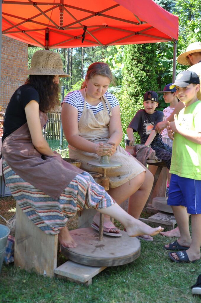 Kobieta wspólnie z dziewczynką tworzą ceramikę na kole garncarskim, wokoło stoi grupa dzieci.
