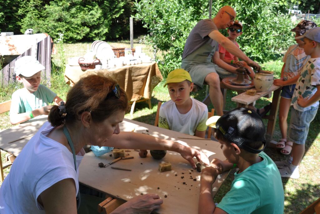 Dzieci przy stole z kobietą, wszyscy zaangażowani wspólne wykonywanie przedmiotów w glinie, w tle mężczyzna i trzech chłopców przy kole garncarskim.