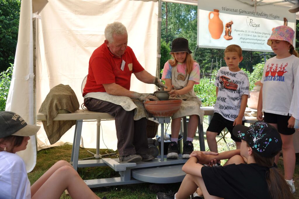 Mężczyzna i dziewczynka wspólnie tworzą ceramikę na kole garncarskim, wokół stoją i siedzą dzieci.