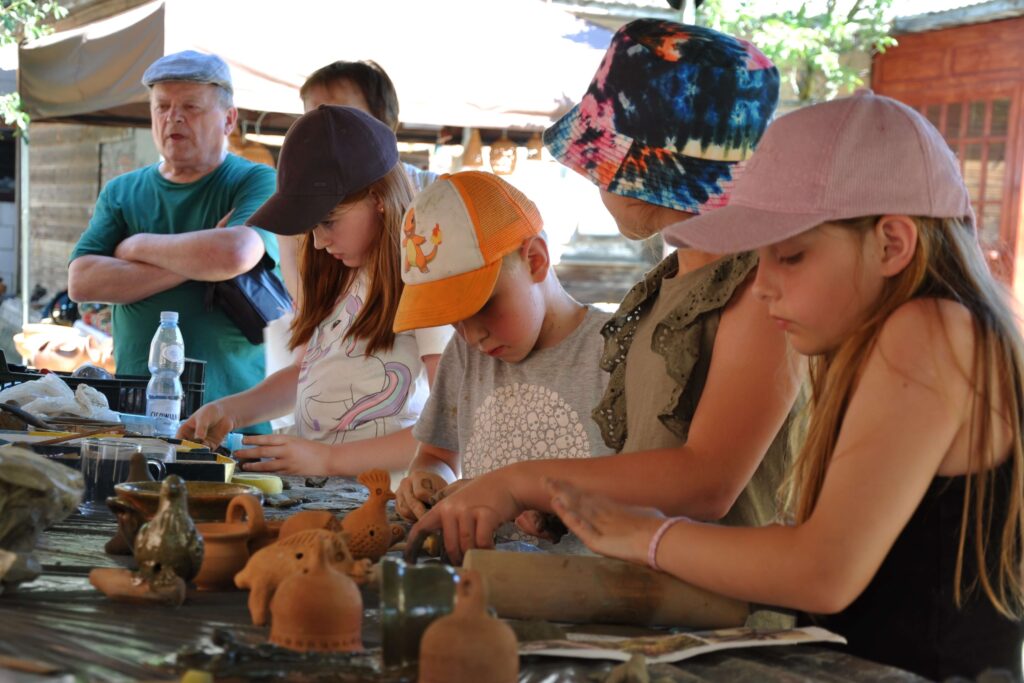 Grupa dzieci przy stole wykonuje przedmioty z gliny, przygląda się dorosły mężczyzna.