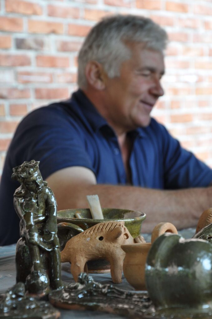 Mężczyzna siedzi przy stole, na którym znajdują się różne wyroby ceramiczne, rzeźby, figurki.