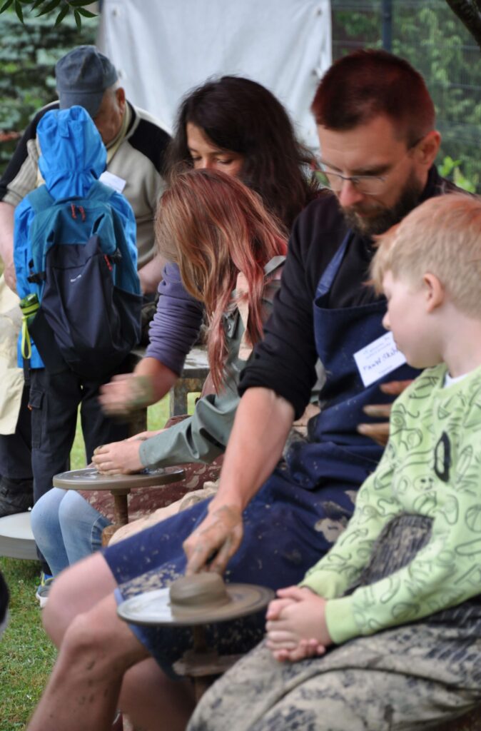 Grupa ludzi, dorośli i dzieci pracują nad ceramiką na kołach garncarskich.