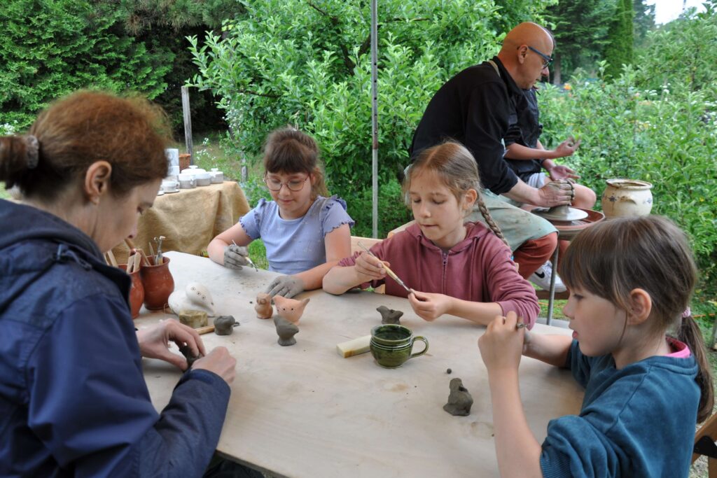 Dzieci przy stole z kobietą, wszyscy zaangażowani wspólne wykonywanie przedmiotów w glinie, w tle mężczyzna pracujący na kole garncarskim.