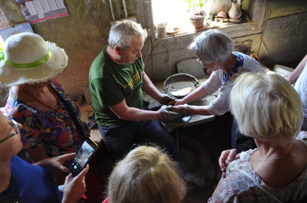 Dwoje dorosłych ludzi pracują wspólnie przy kole garncarskim, wokoło stoją i obserwują inne osoby.