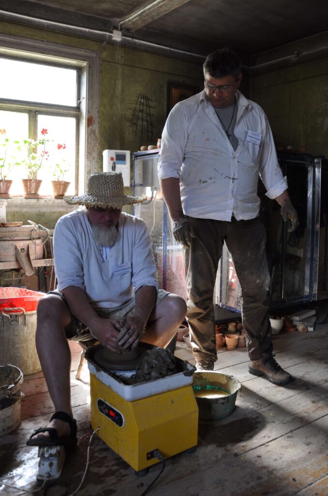 Mężczyzna pracuje nad ceramiką na kole garncarskim, obok niego stoi drugi mężczyzna i obserwuje.