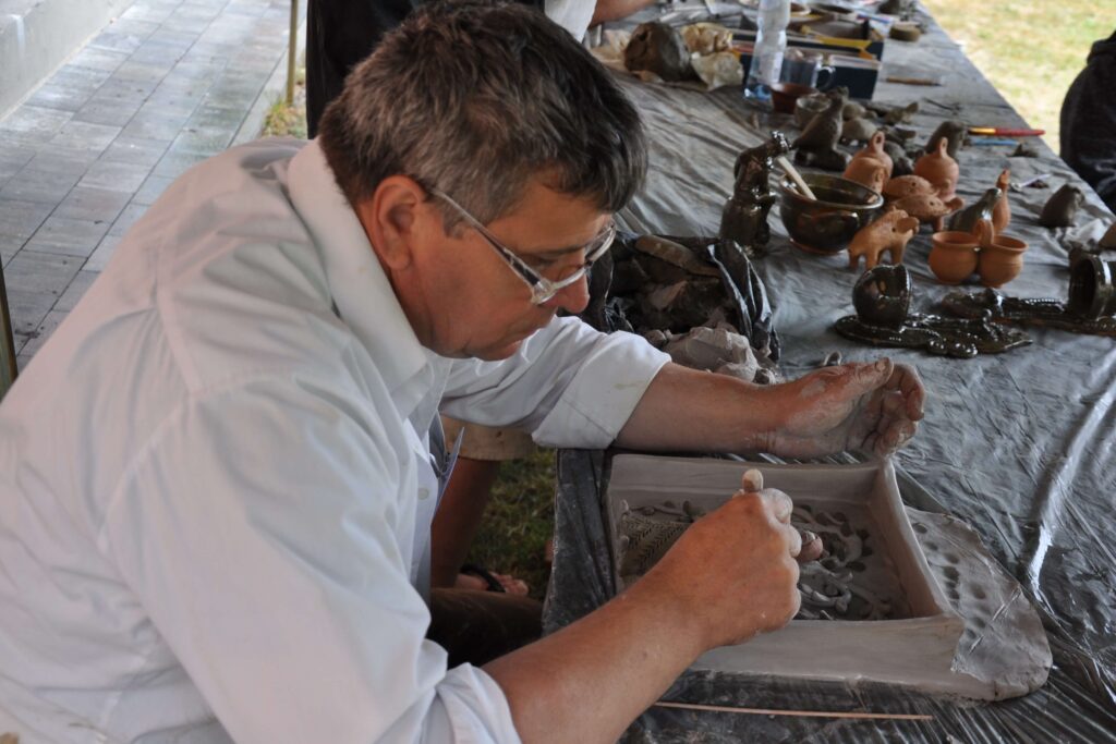 Mężczyzna w białej koszuli lepi kapliczkę z gliny przy stole.