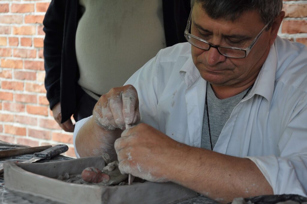 Mężczyzna w białej koszuli lepi kapliczkę z gliny przy stole.