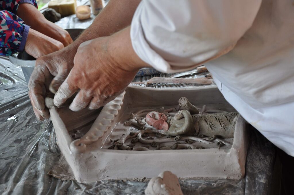 Ręce mężczyzny w białej koszuli wykonujące kapliczkę z gliny na stole.