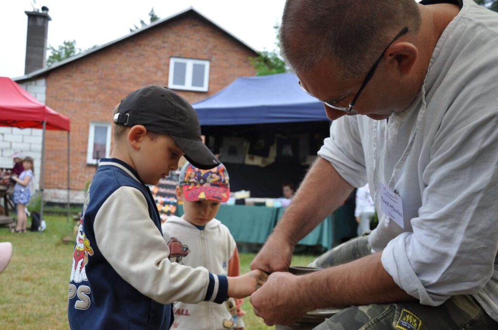 Mężczyzna i chłopiec pracują nad ceramiką na kole garncarskim, obok stoi drugi chłopiec i przygląda się ich pracy.