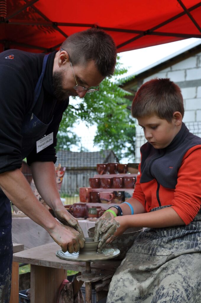 Mężczyzna i chłopiec wspólnie lepią ceramikę siedząc przy kole garncarskim.