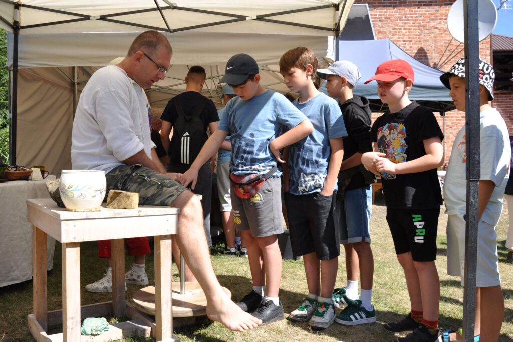 Mężczyzna siedzi przy kole garncarskim pod namiotem, przy nim stoi pięciu chłopców w kolejce.