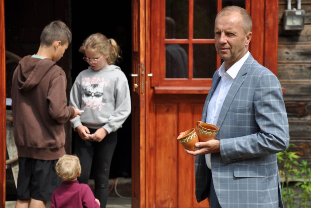 Mężczyzna w garniturze trzyma dwa malowane kubki gliniane, w tle trójka dzieci stoi w drzwiach starego domu.