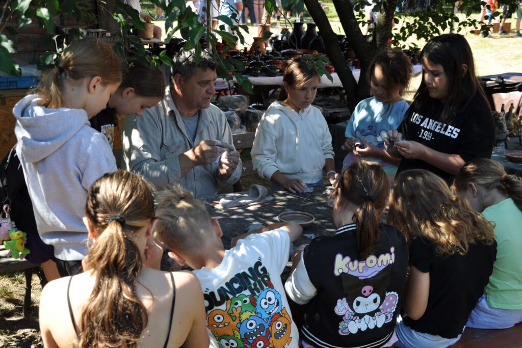 Grupa dzieci siedzi przy stole z mężczyzną, wszyscy zaangażowani w rozmowę i lepienie z gliny.