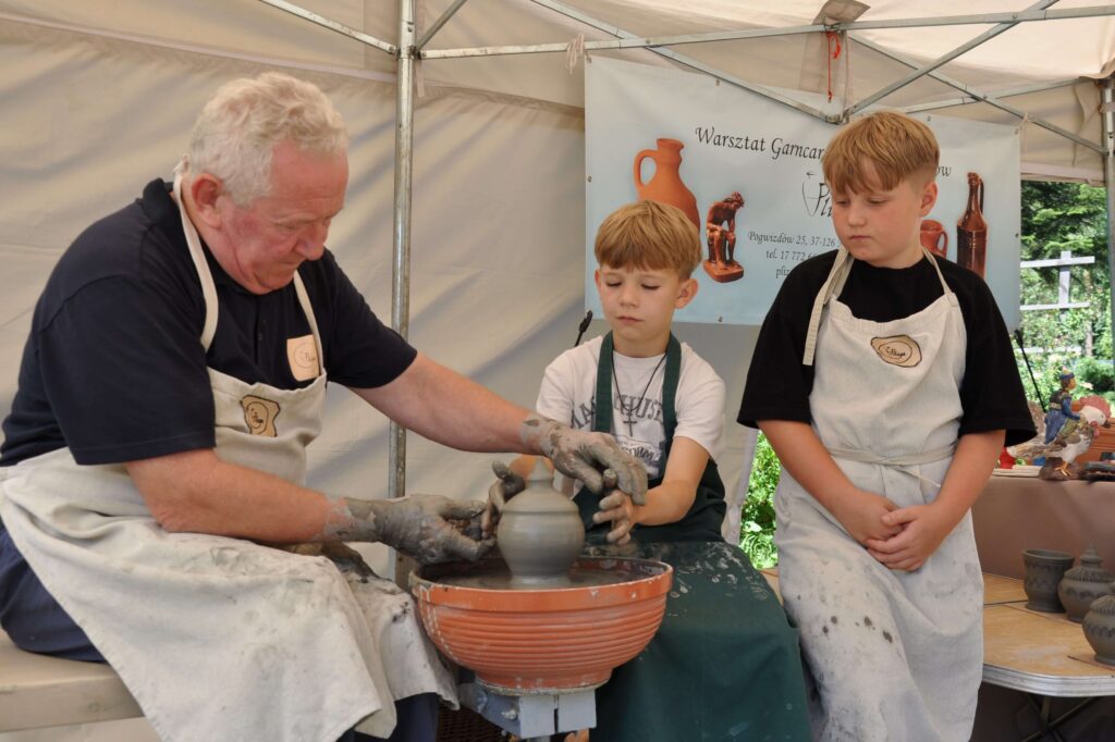 Mężczyzna i chłopiec wspólnie lepią ceramikę siedząc przy kole garncarskim, obok nich siedzi drugi chłopiec i obserwuje ich pracę.