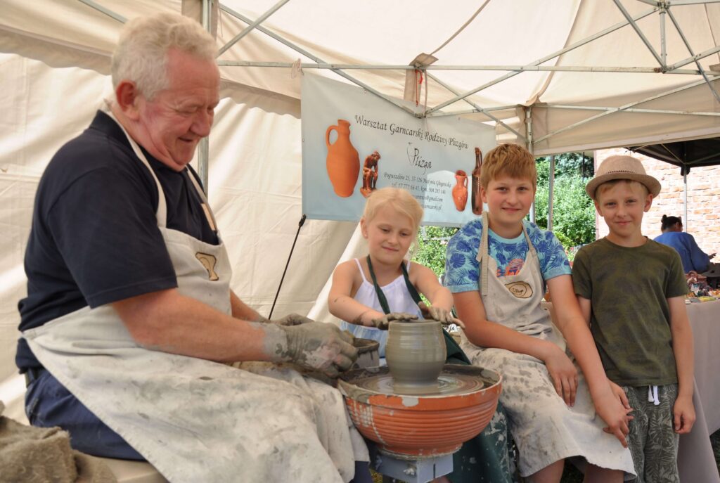 Mężczyzna i i mała dziewczynka wspólnie lepią ceramikę siedząc przy kole garncarskim, obok dwóch chłopców obserwuje ich pracę.