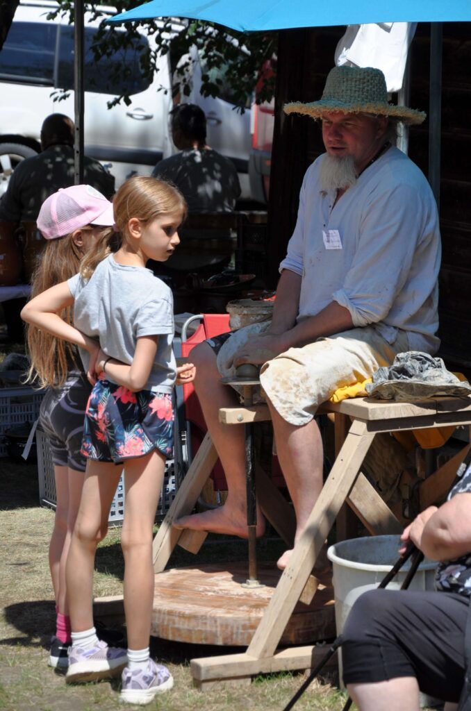 Mężczyzna lepi ceramikę siedząc przy kole garncarskim, obok stoją dwie dziewczynki obserwując jego pracę.