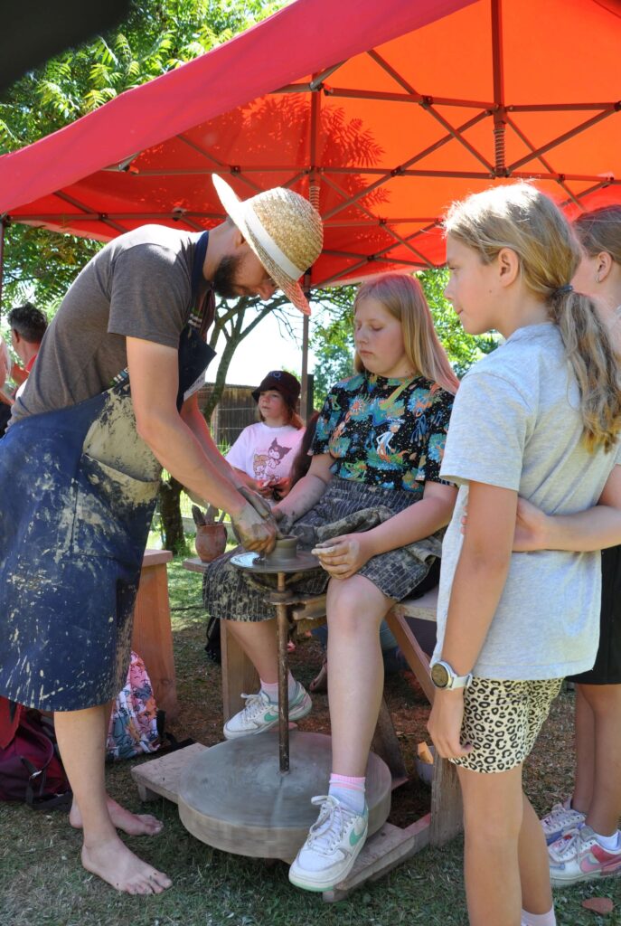 Mężczyzna i dziewczynka wspólnie lepią ceramikę siedząc przy kole garncarskim, wokoło stoi grupa dzieci.