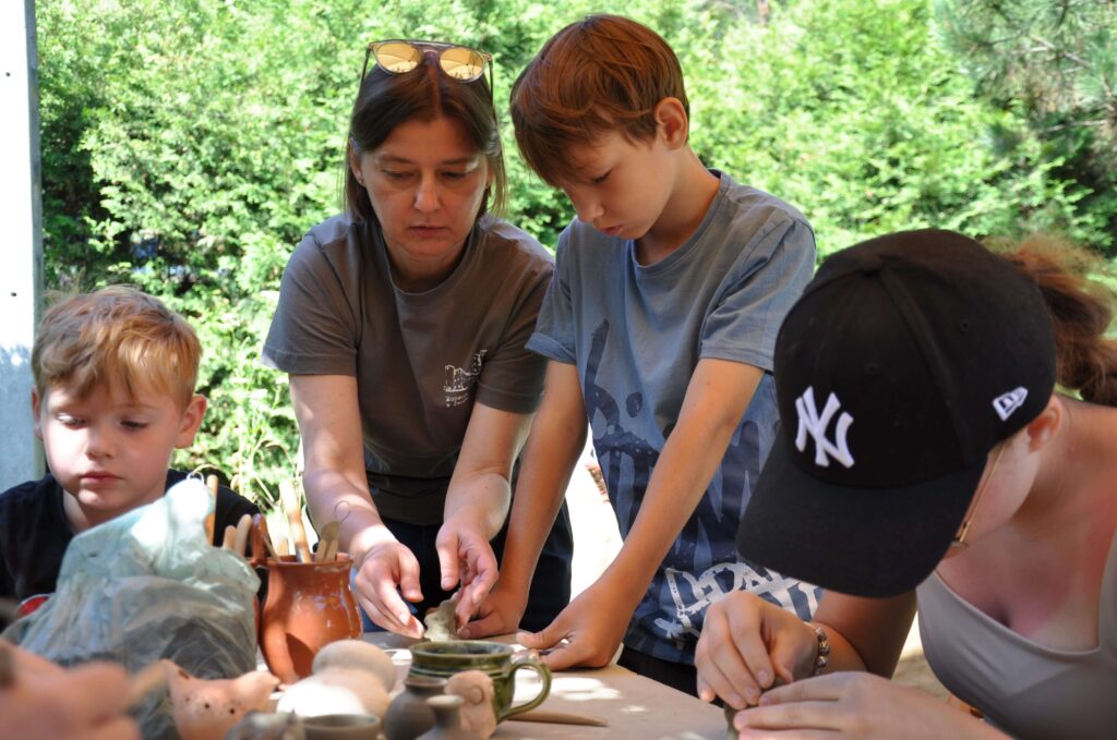 Kobieta z dziećmi siedzi przy stole, wszyscy skupieni na twórczej pracy z gliną.