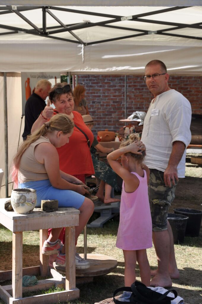 Dziewczynka przy kole garncarskim tworzy naczynie gliniane, obok niej stoi kobieta z uniesionym w górę kciukiem, mężczyzna w białej koszuli i mała dziewczynka.