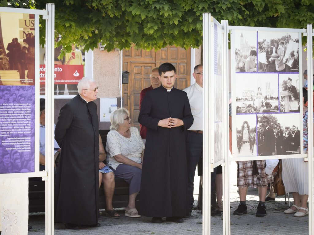 Dwóch księży stoi pod drzewem przez planszami ze zdjęciami, między nimi siedzi na ławce starsza kobieta.