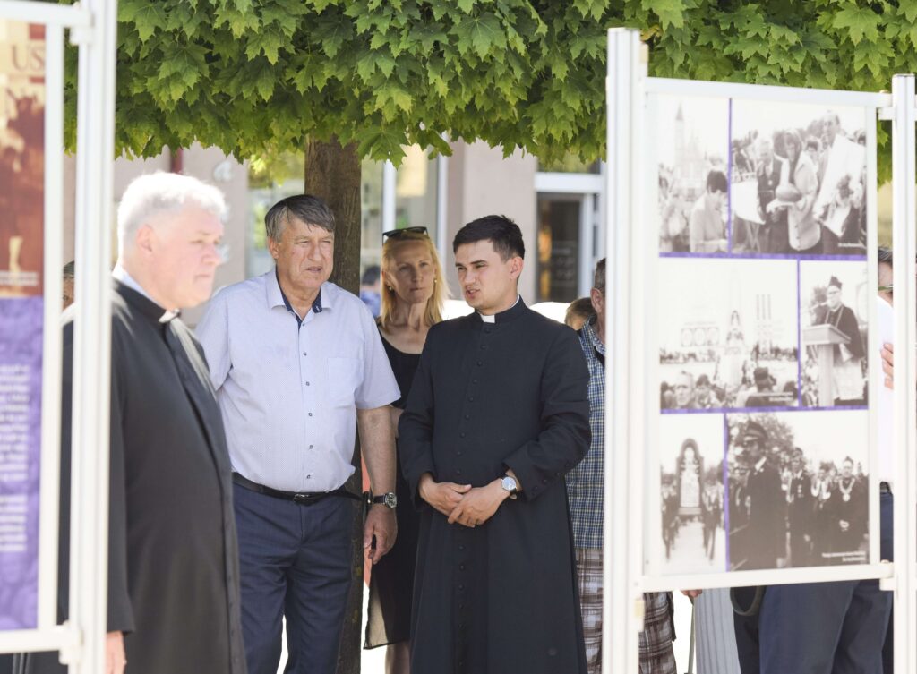 Dwóch księży oraz mężczyzna i kobieta stoją pod drzewem przez planszami ze zdjęciami.
