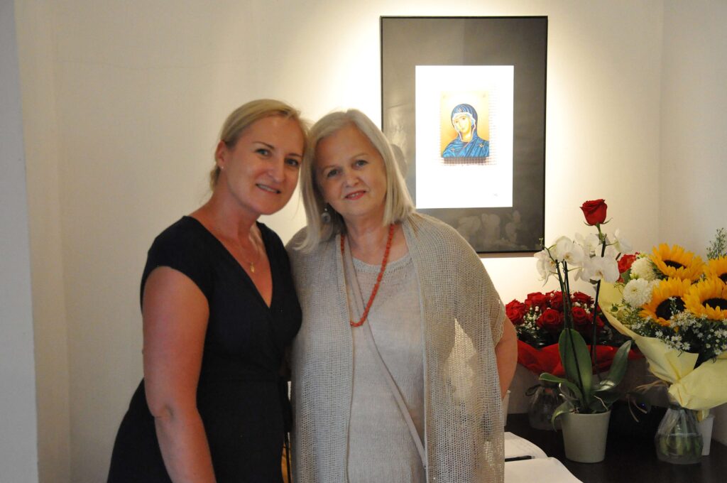 Dwie kobiety pozują do zdjęcia na tle ściany z zawieszonym na niej plakatem z wizerunkiem Matki Bożej.