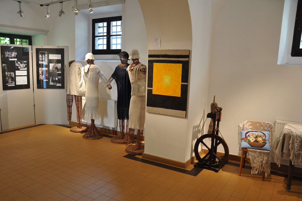 Sala wystawowa z zawieszonymi na ścianie zdjęciami, kolorowym obrazem, czterema manekinami ubranymi w różne stroje i kołowrotkiem.