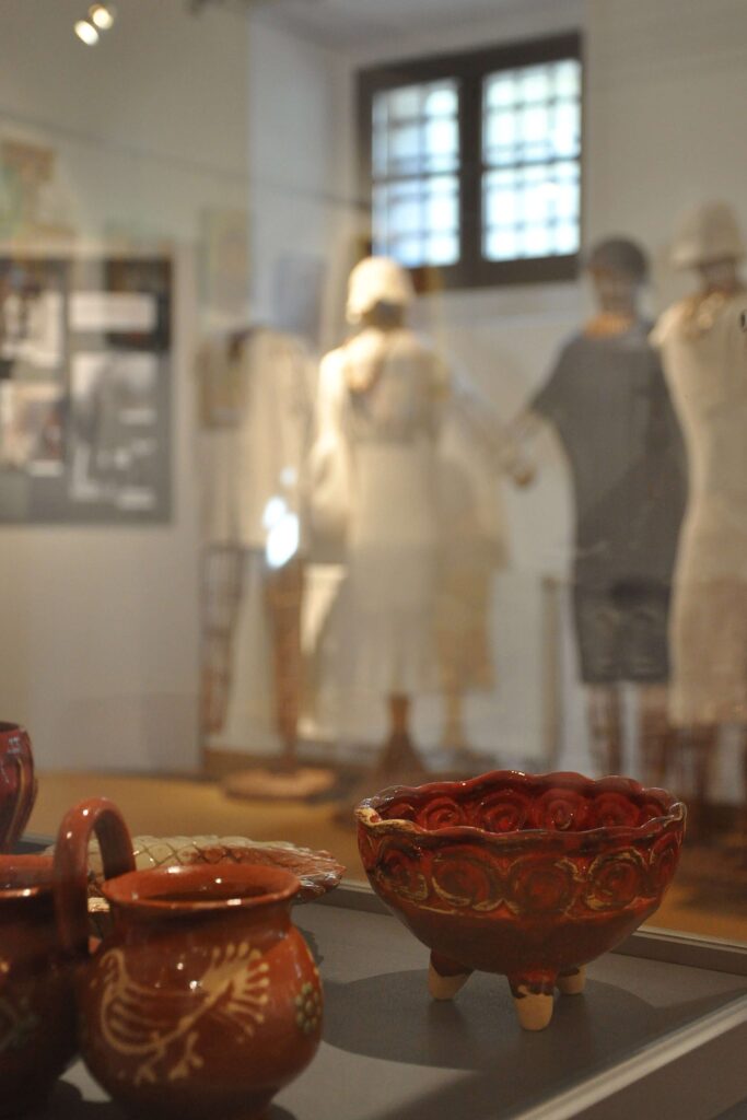 Ceramika gliniana w szklanej gablocie na tle ściany z zawieszonymi zdjęciami i stojącymi manekinami ubranymi w różne stroje.