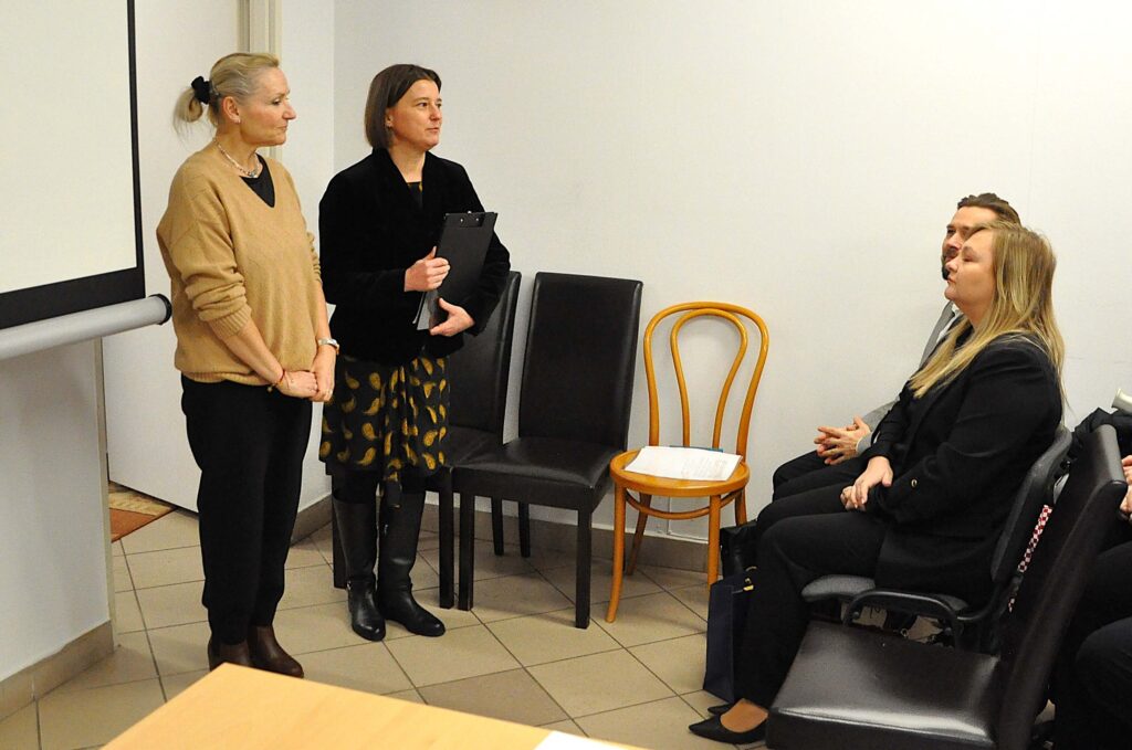 Cztery osoby w pomieszczeniu, dwie stojące kobiety po lewej, dwie siedzące osoby po prawej, krzesła w tle, jasne ściany, podłoga z beżowych płytek.