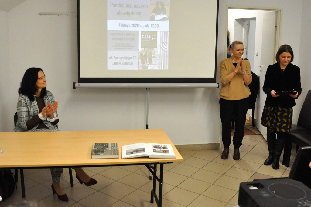 Trzy kobiety w pomieszczeniu, jedna siedzi przy stole z książkami, dwie stoją obok otwartych drzwi, na ścianie ekran z projekcją tekstu i zdjęcia.