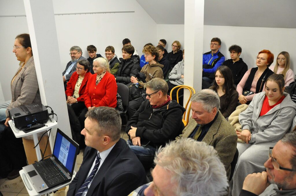 Grupa osób w różnym wieku siedzi w pomieszczeniu z białymi ścianami, obok stoi kobieta ubrana w jasną marynarkę, obok niej projektor i laptop na stoliku.