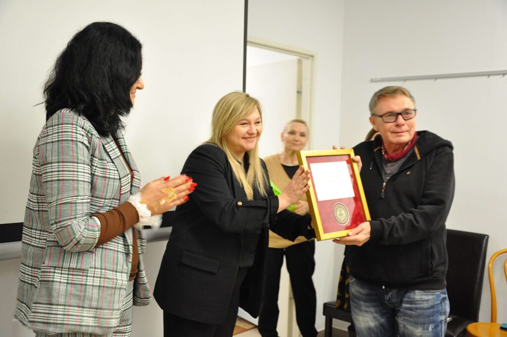 Cztery osoby w pomieszczeniu, kobieta w czarnej marynarce podaje mężczyźnie w okularach ramkę z dyplomem, druga kobieta z czarnymi włosami klaszcze, trzecia stoi w tle.