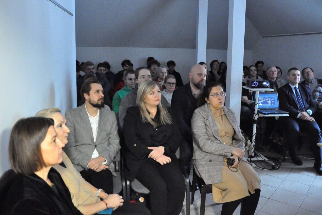 Grupa ludzi siedzących w pomieszczeniu, większość w ciemnych ubraniach, z laptopem i projektorem na stoliku po prawej stronie.