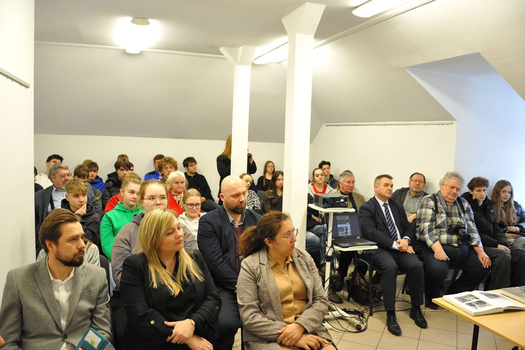 Grupa ludzi siedzących w jasnym pomieszczeniu, wśród nich mężczyźni i kobiety w różnym wieku, w casualowych i formalnych ubraniach, z projektorem i laptopem na stoliku.