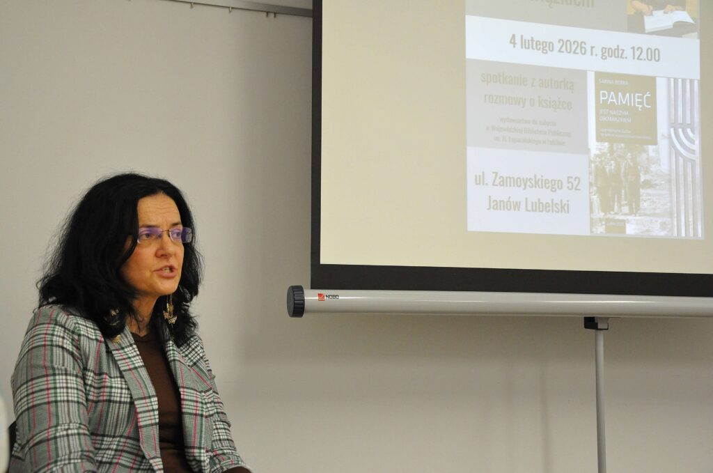 Kobieta w okularach i marynarce w kratę obok ekranu z napisem o spotkaniu autorskim i okładką książki na jasnym tle.