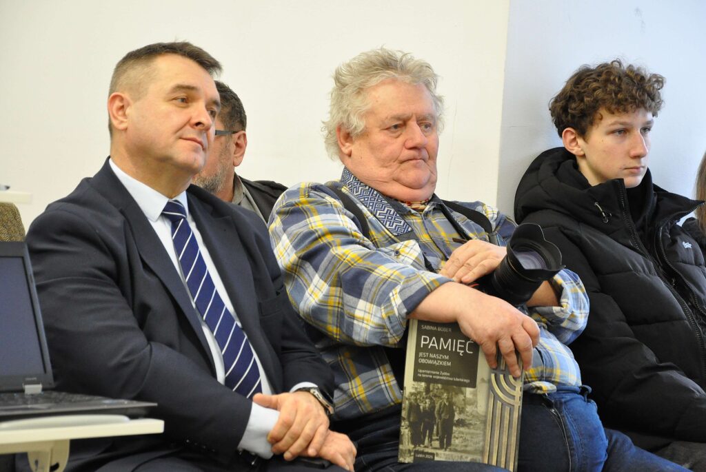 Trzech mężczyzn siedzi na tle jasnej ściany, jeden w garniturze z krawatem, drugi w koszuli w kratę trzyma książkę i aparat, trzeci w czarnej kurtce puchowej.