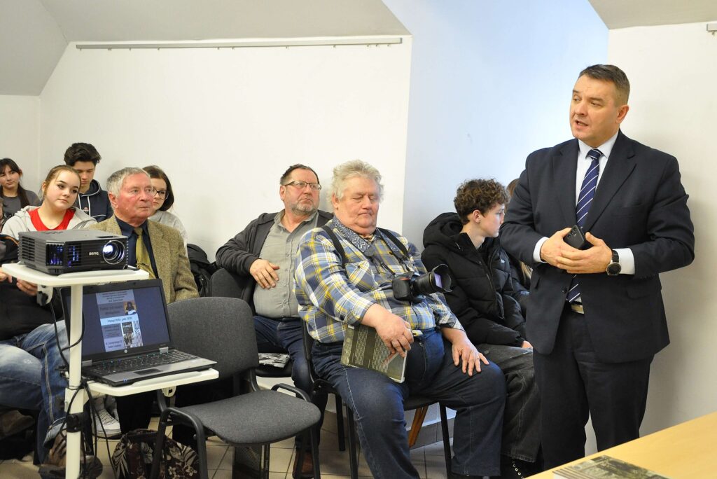 Grupa osób siedzących i słuchających mężczyzny w garniturze przemawiającego obok białej ściany, obok laptop i projektor na stoliku.
