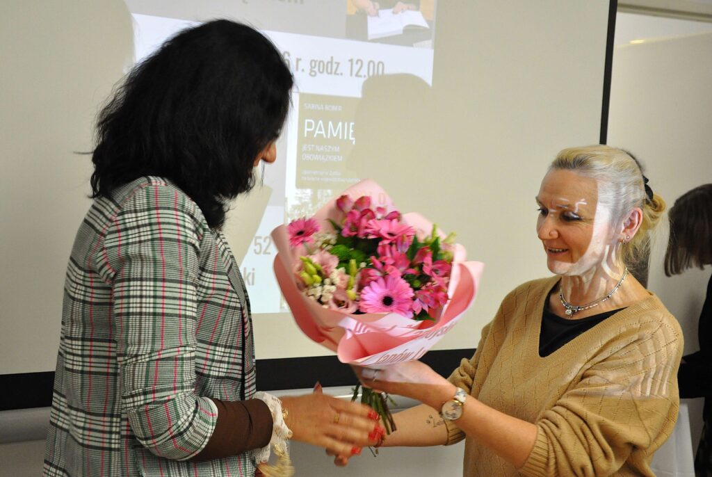 Dwie kobiety, jedna z krótkimi blond włosami w beżowym swetrze przekazuje bukiet różowych kwiatów kobiecie z ciemnymi włosami w kratowanej marynarce.