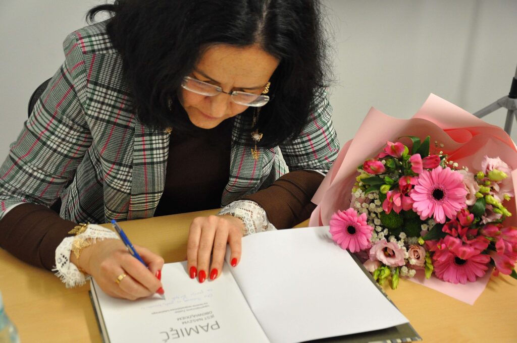 Kobieta z ciemnymi włosami i okularami, w marynarce w kratę, podpisuje książkę, obok leży bukiet różowych kwiatów na jasnym stole.