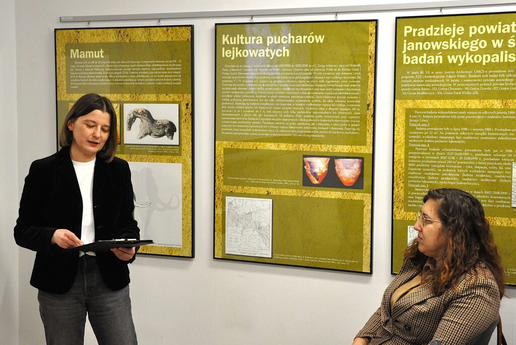 Dwie kobiety, jedna stoi z teczką w rękach ubrana w czarną marynarkę i białą koszulę, druga siedzi w kraciastej marynarce i okularach, za nimi żółte plansze z tekstem na ścianie.