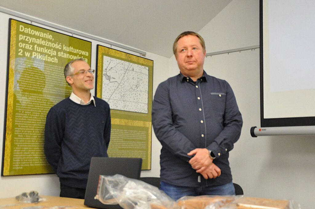 Dwaj mężczyźni stojący obok stołu z laptopem i przezroczystą torbą, za nimi zielone plansze z tekstem i mapą na ścianie, po prawej ekran projekcyjny.