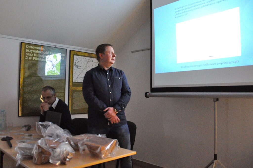 Mężczyzna w ciemnej koszuli stoi obok stołu z plastikowymi pakunkami, za nim siedzi inny mężczyzna, na ścianie plansze z mapami i tekstem, po prawej ekran projekcyjny.