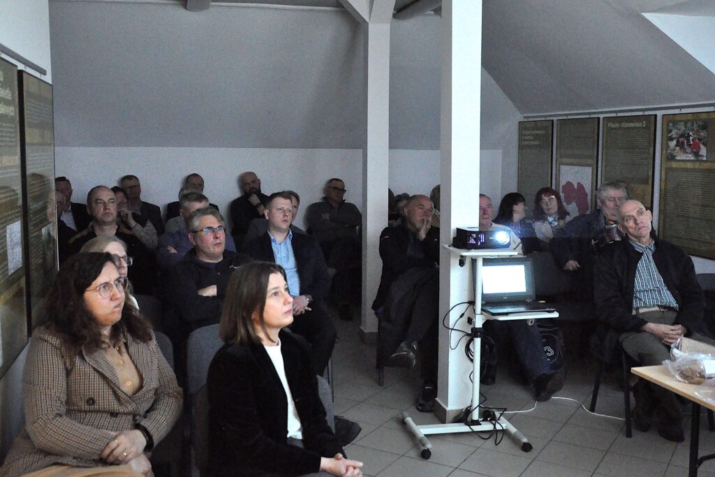 Grupa dorosłych osób siedzących w sali, z przodu laptop i rzutnik na stojaku, na ścianach plansze informacyjne, wnętrze z białymi ścianami i szarym sufitem.