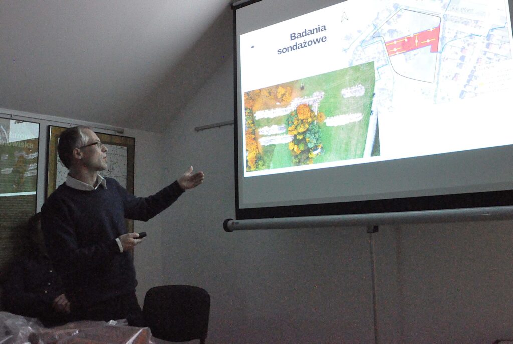 Mężczyzna w ciemnym swetrze i jasnej koszuli wskazuje na ekran z mapą i napisem "Badania sondażowe" w ciemnym pomieszczeniu.