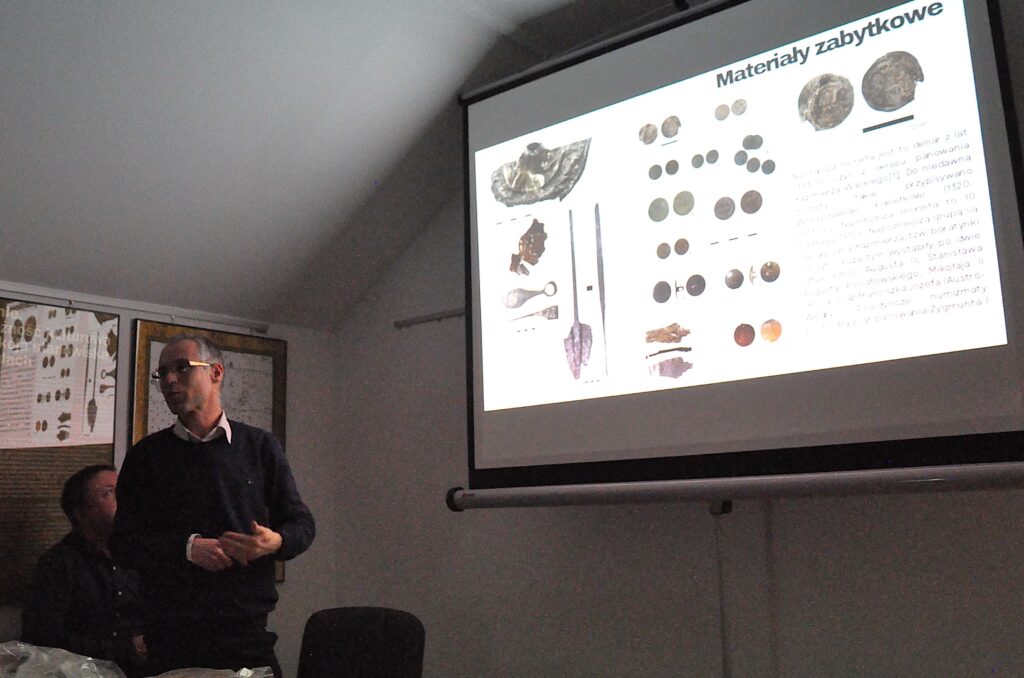 Mężczyzna w ciemnym swetrze i białej koszuli stoi obok ekranu z projekcją pokazującą zabytkowe monety i narzędzia na białym tle w ciemnym pomieszczeniu.
