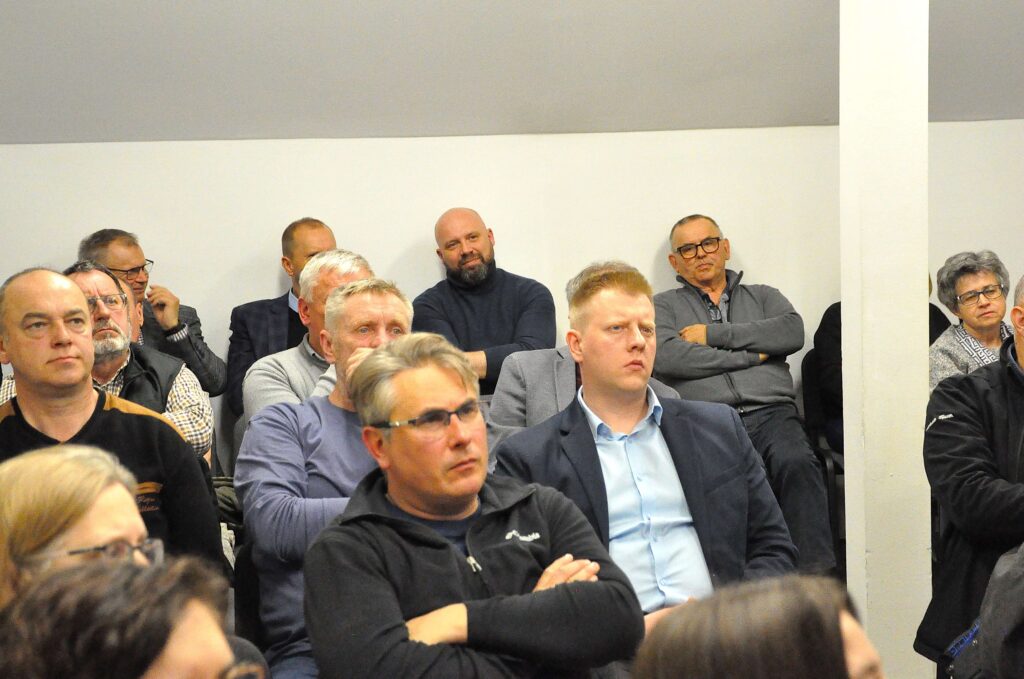 Grupa osób siedzących na krzesłach w pomieszczeniu z białą ścianą i słupem, większość mężczyzn w średnim wieku, ubrani w casualowe i formalne ubrania.