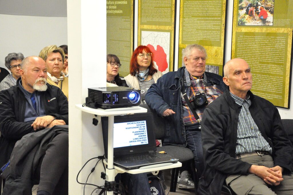 Grupa dorosłych osób siedzi na krzesłach, na pierwszym planie projektor i laptop z wyświetlanym tekstem, w tle zielone tablice informacyjne na ścianie.